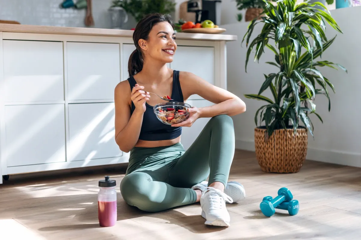 Smiling woman sitting on the floor eating a healthy bowl of fruit and cereal after a workout.