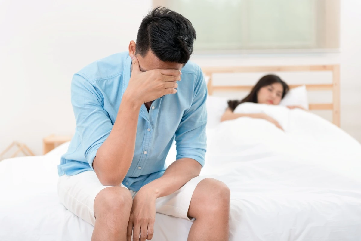 Stressed man sitting on the edge of the bed covering his face while a woman lies in the background.