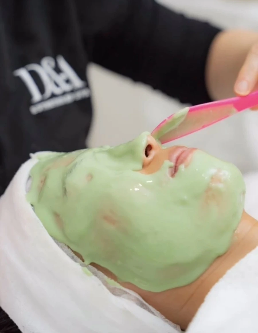 Professional esthetician applying a soothing green facial mask with a spatula during a spa treatment.